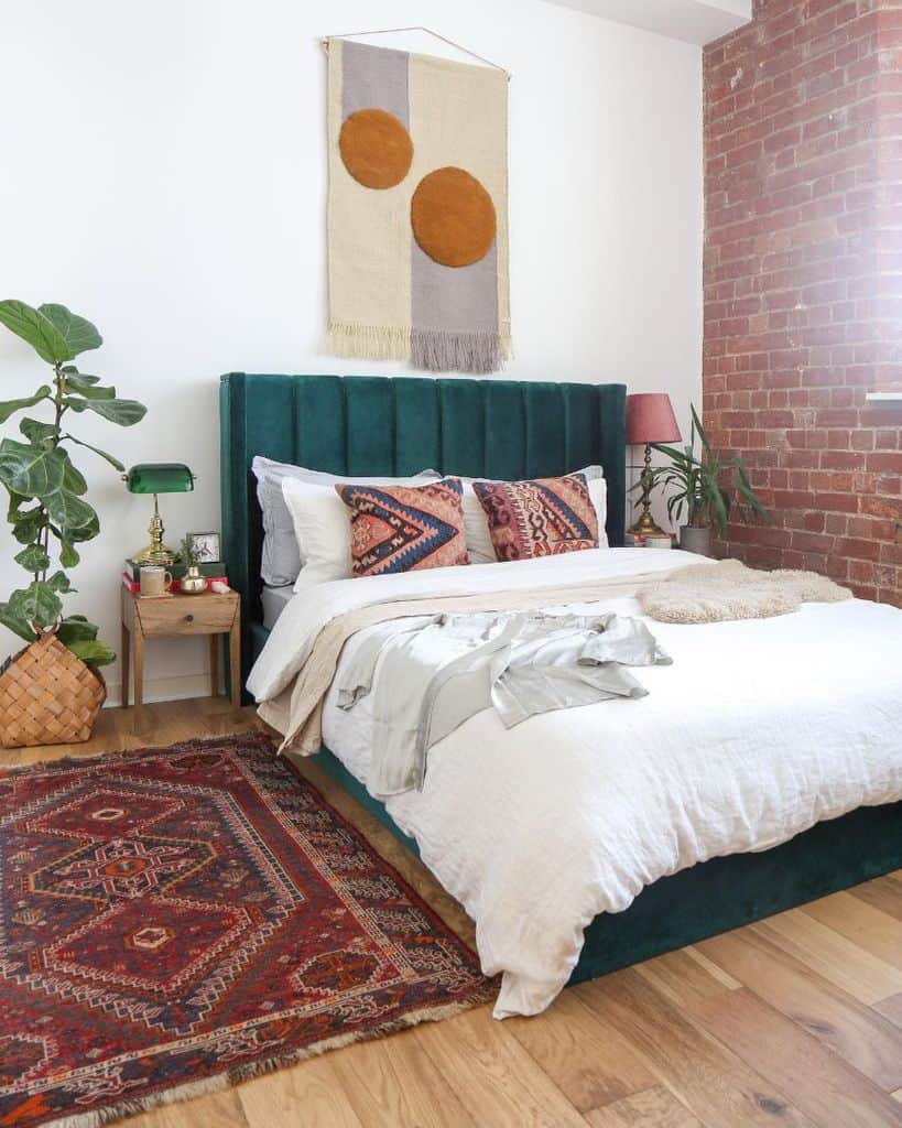 Boho bedroom with green velvet headboard, brick accent wall, patterned rug, and colorful pillows.