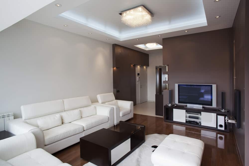 Modern living room with white sofas, brown wood furniture, TV, and recessed lighting on a white and brown ceiling