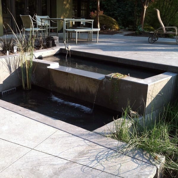 Concrete waterfall feature flowing into a small pond, surrounded by patio seating and modern landscaping.