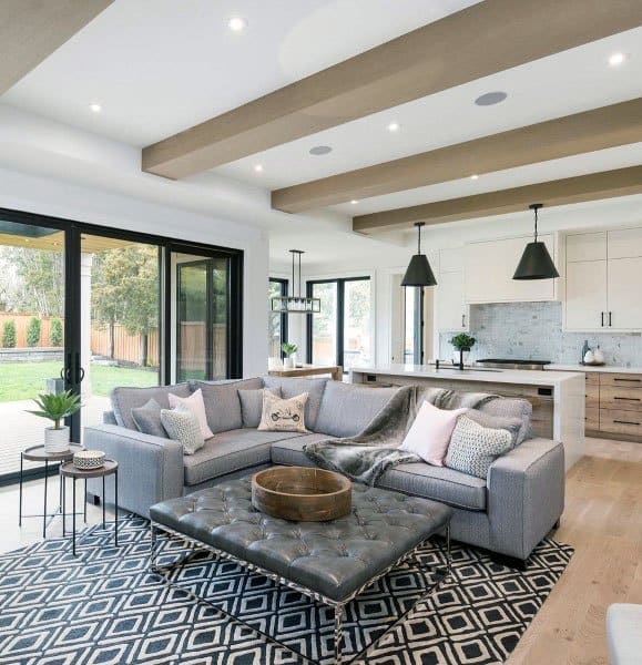 Modern living room with gray sectional, patterned rug, tufted coffee table, and open kitchen featuring pendant lights