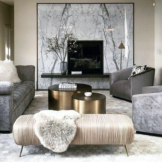 Elegant living room with a marble fireplace, gray sofas, a textured ottoman, and metallic coffee tables