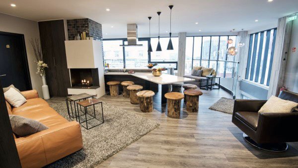 Modern living room and kitchen with leather couch, stools, a cozy fireplace, large windows, neutral tones, and wooden accents