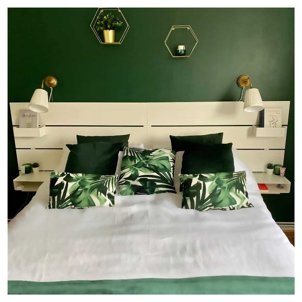 A stylish bedroom with a white bed, green pillows, leaf-patterned cushions, and a dark green accent wall with decor