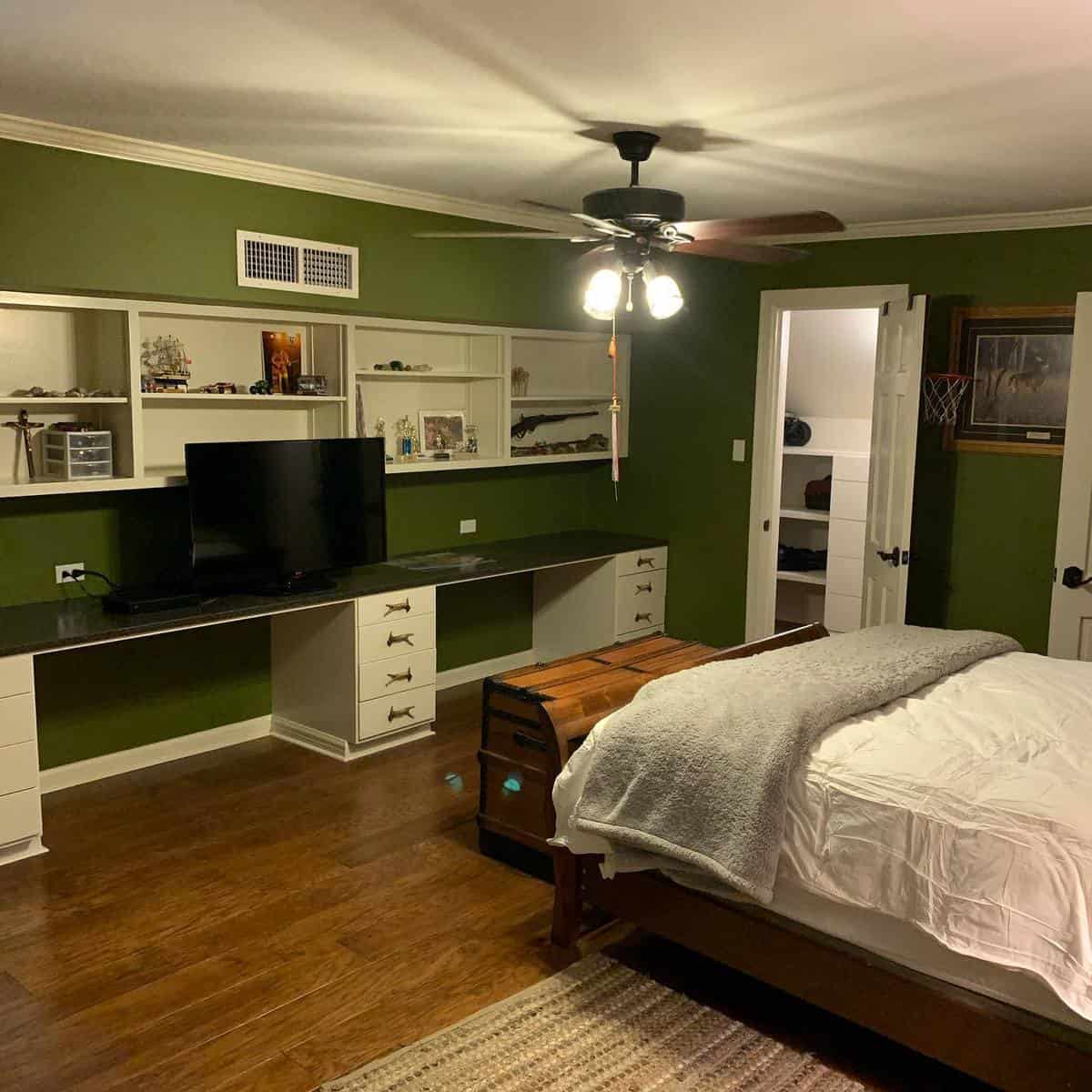 Bedroom with green walls, ceiling fan, TV on a desk, wooden chest at bed's foot, and open door to another room