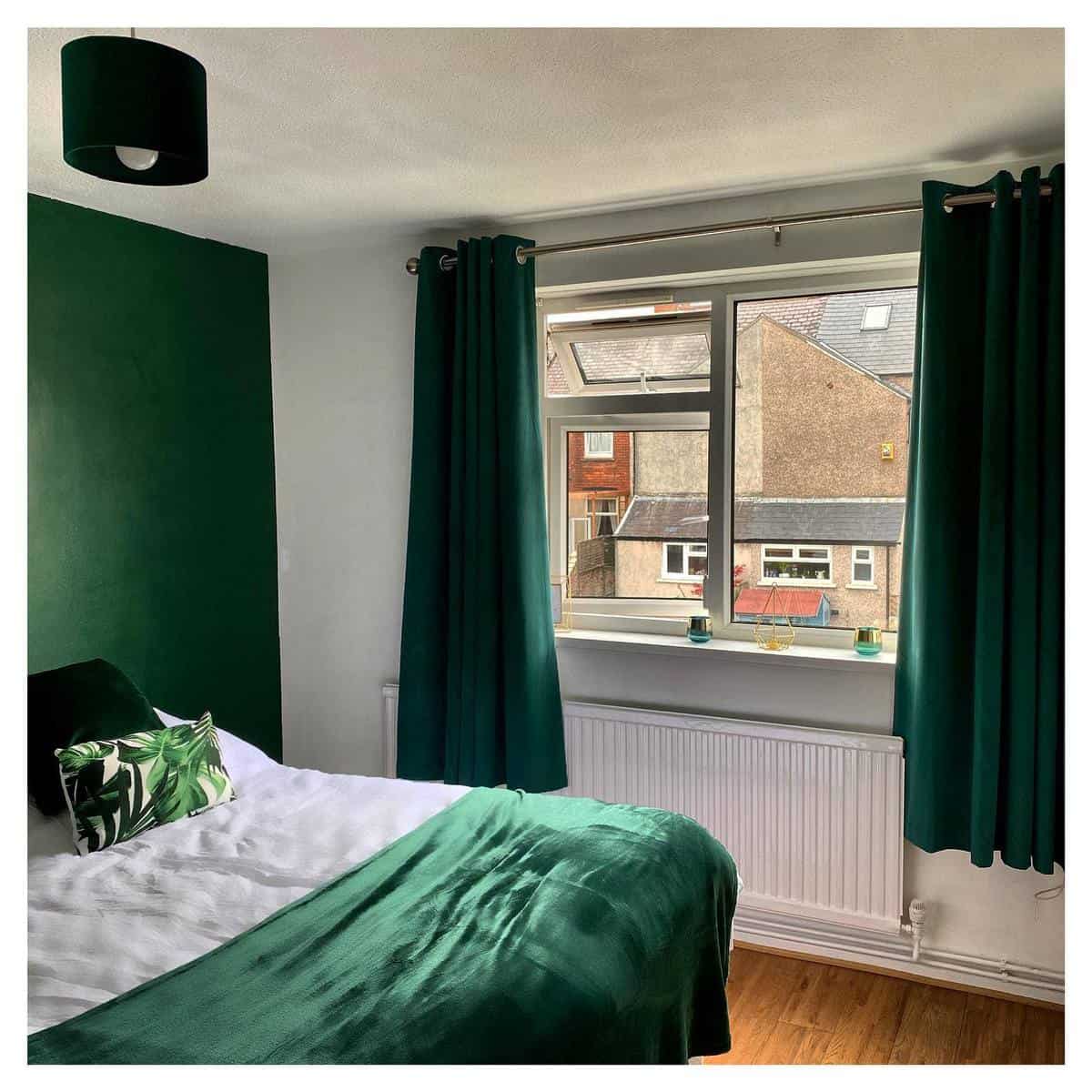 Cozy bedroom with green accents, including curtains, pillows, and bedspread, featuring a view of nearby buildings