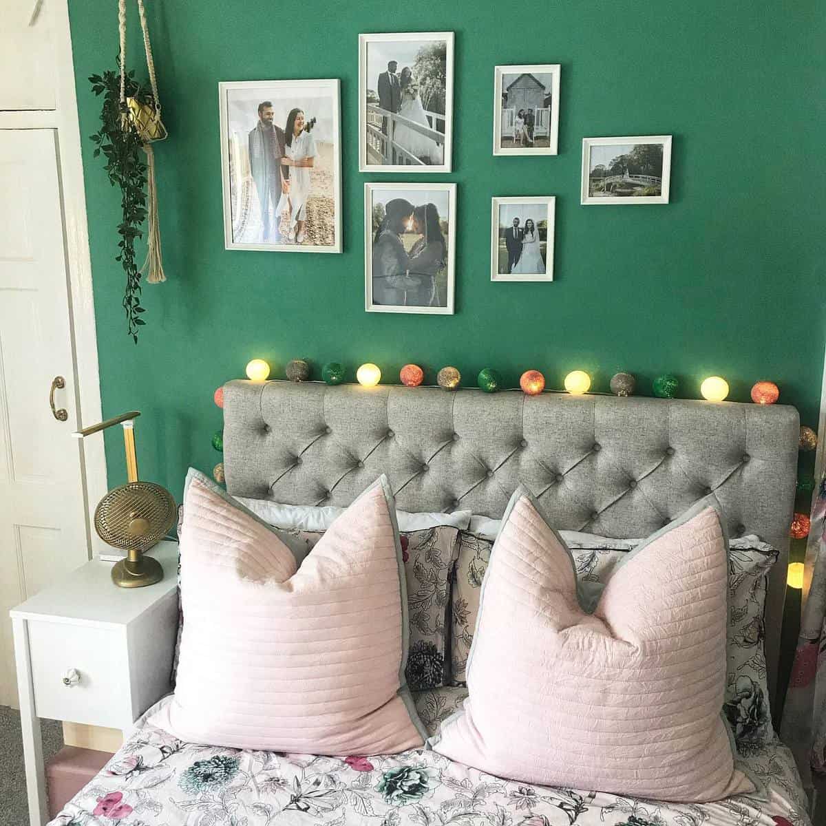 Bedroom with a gray tufted headboard, pink pillows, green wall with photos, and string lights above the bed