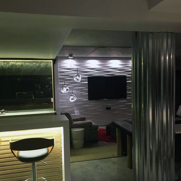 Modern man cave with illuminated wet bar, corrugated metal column, and wall-mounted TV in lounge area.