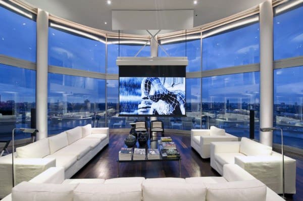 Sleek living room with curved glass walls, white sofas, a large screen, and cityscape views