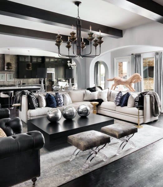 Modern living room with a white sofa, black table, chandeliers, and silver decor; kitchen in the background