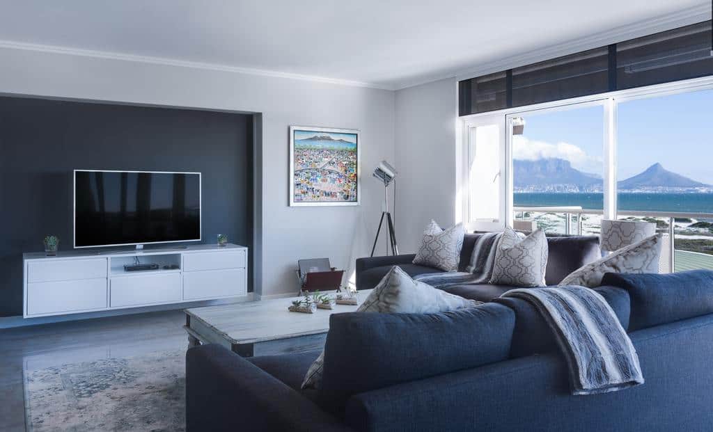 Modern minimalist living room with navy blue sofas, a flat-screen TV, and large windows offering stunning ocean and mountain views