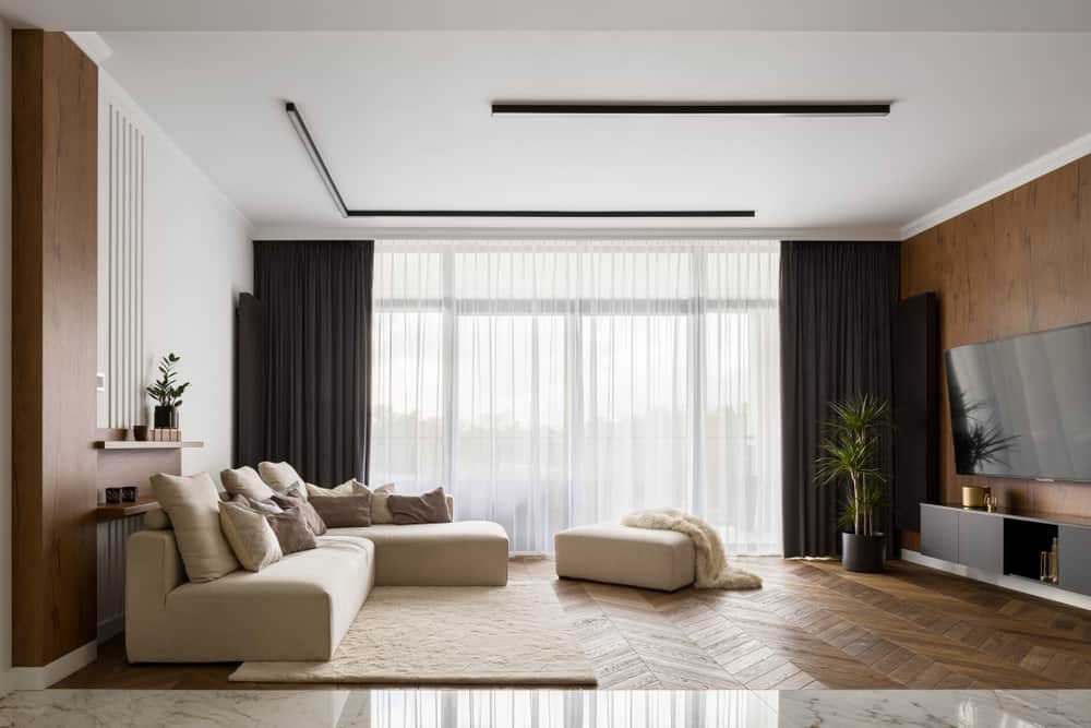 Modern living room with a cream sofa, wooden accents, floor-to-ceiling curtains, and herringbone parquet flooring