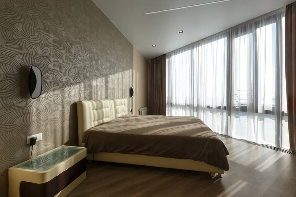 Modern bedroom with patterned wallpaper, large windows, and minimalist beige furnishings.