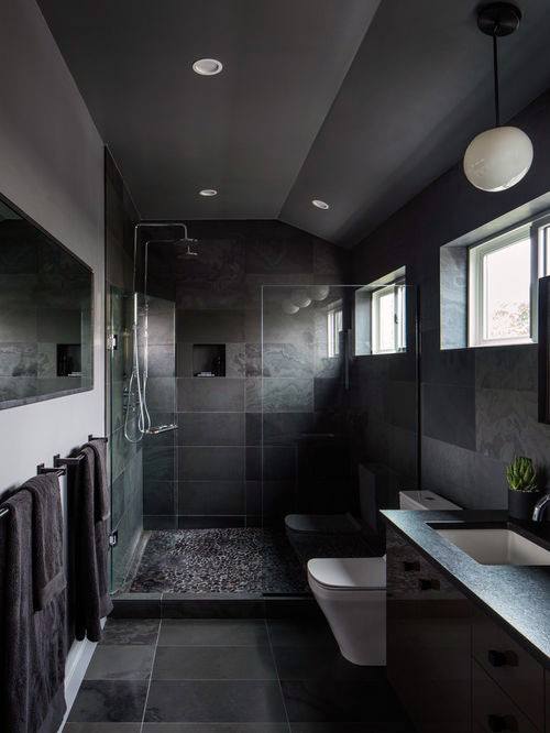 Modern bathroom with dark tiles, glass shower, pebble floor, and minimalist lighting.