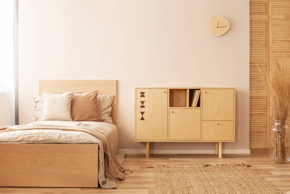 Minimalist bedroom with wooden bed, beige bedding, a light wood cabinet, woven rug, and wall clock