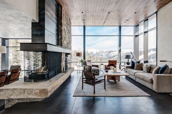 A rustic living room with a modern design featuring a stone fireplace, large windows offering stunning mountain views, and sleek, comfortable furniture, creating a spacious and inviting atmosphere