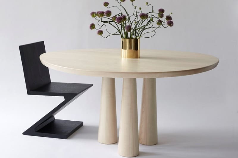 Round wooden table with sculptural legs, black chair, and floral arrangement centerpiece.