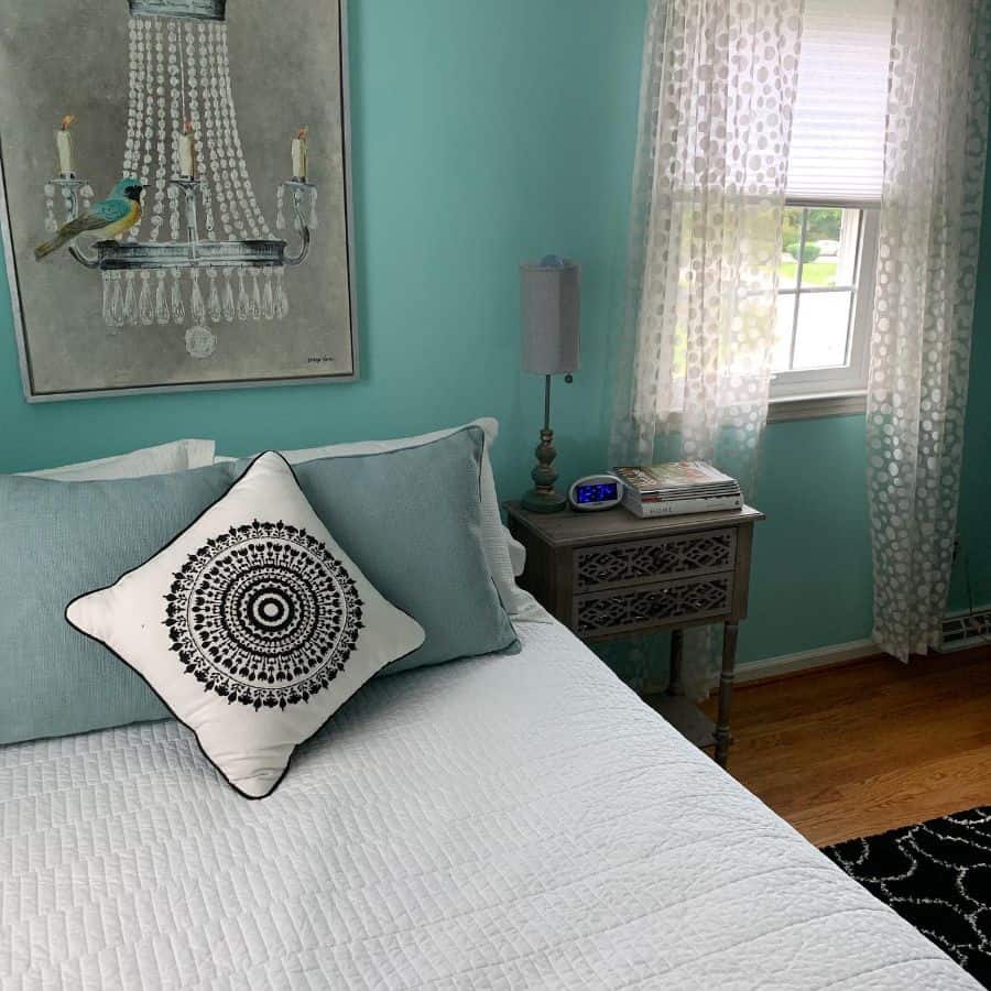 Teal bedroom with patterned pillow, chandelier artwork, and decorative side table.