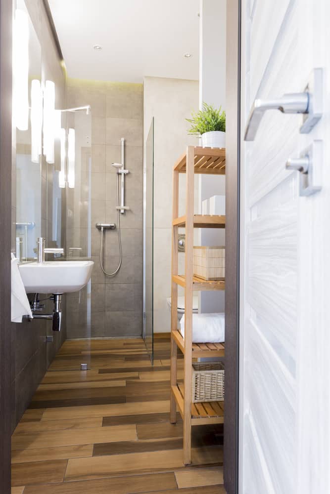 Narrow modern bathroom with gray tiles, wood-look flooring, and wooden shelving unit.