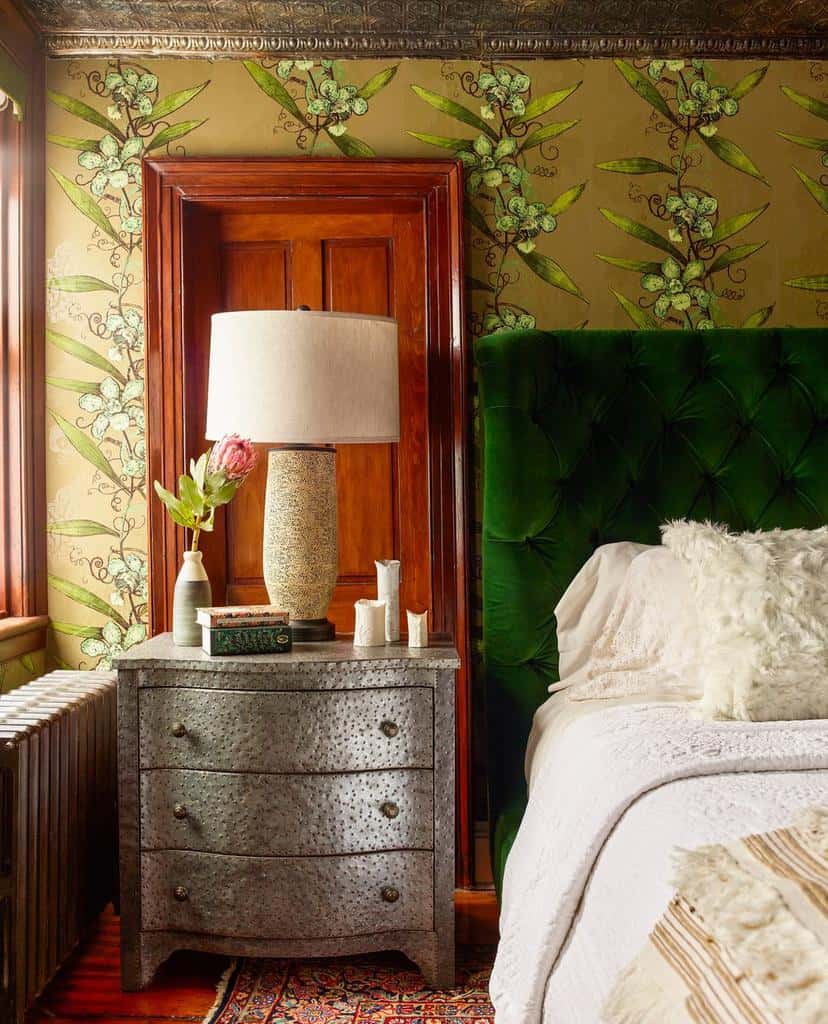 Green velvet headboard with floral wallpaper, wooden door, and textured nightstand.