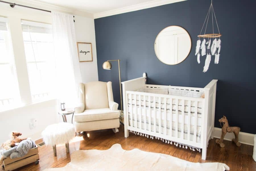 Modern baby room with white crib, deep blue accent wall, and dreamcatcher mobile above hardwood floors.