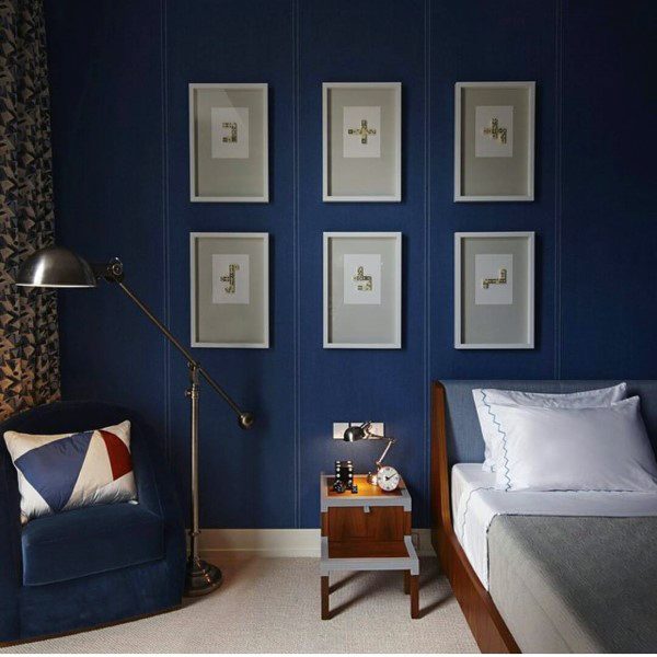 Navy blue bedroom with framed wall art, wooden bed, blue armchair, and floor lamp.