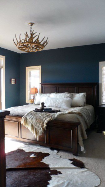 Rustic navy bedroom with wooden bed frame, antler chandelier, and cowhide rug.