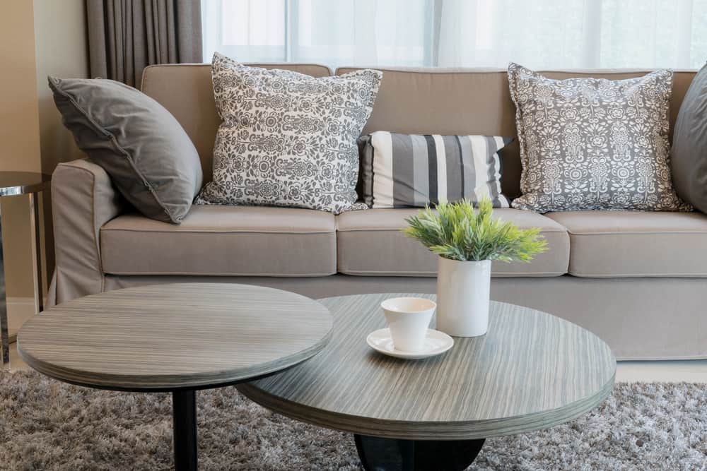 Cozy living room with a beige sofa, patterned pillows, brown round tables, a potted plant, and a cup on a saucer