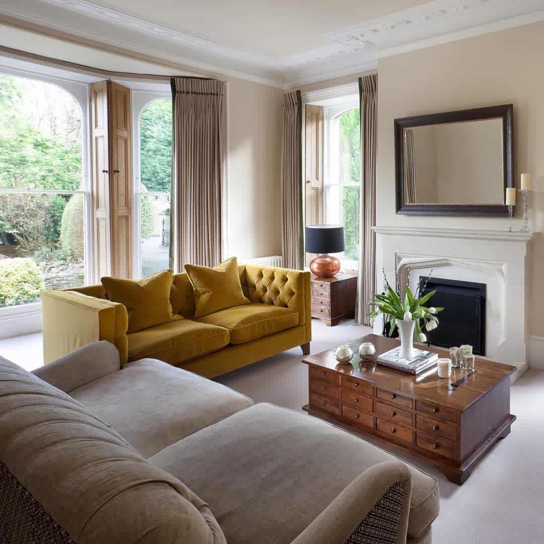 Cozy living room with a yellow sofa, mirror above a white fireplace, and a brown coffee table by large windows