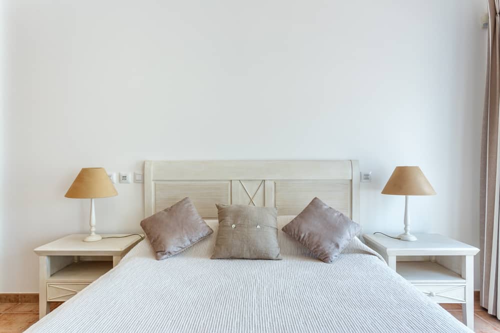 Minimalist bedroom with a white bed, beige pillows, white side tables, and matching lamps against a plain wall