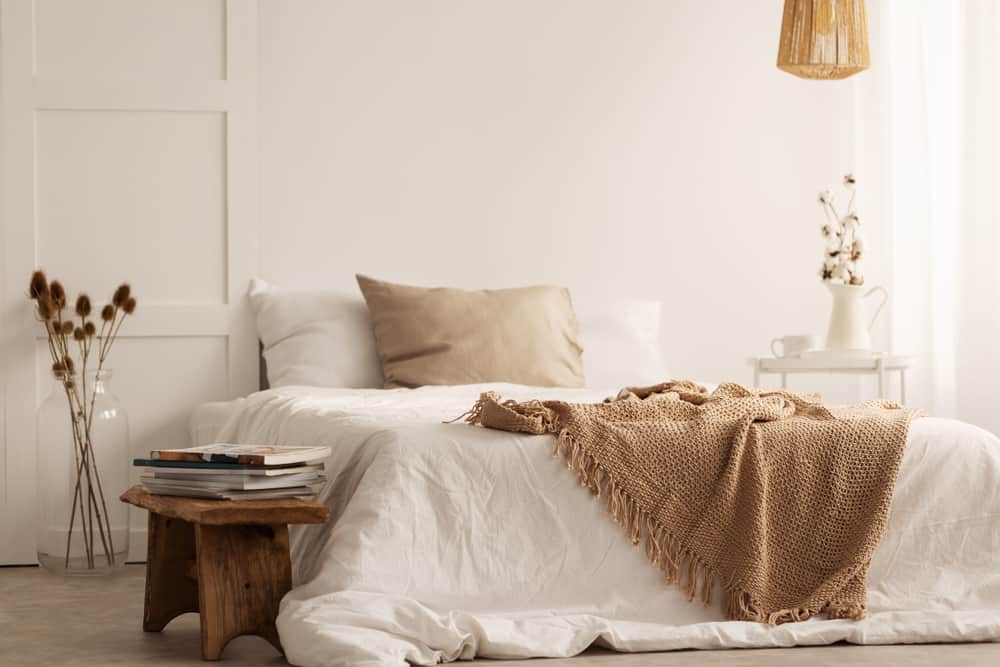 Cozy beige bedroom with a bed, woven blanket, books on a white wooden bench, and dried flowers in a vase