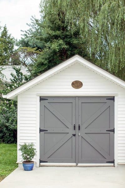 White shed with grey farmhouse-style double doors and a small potted plant on a paved path.