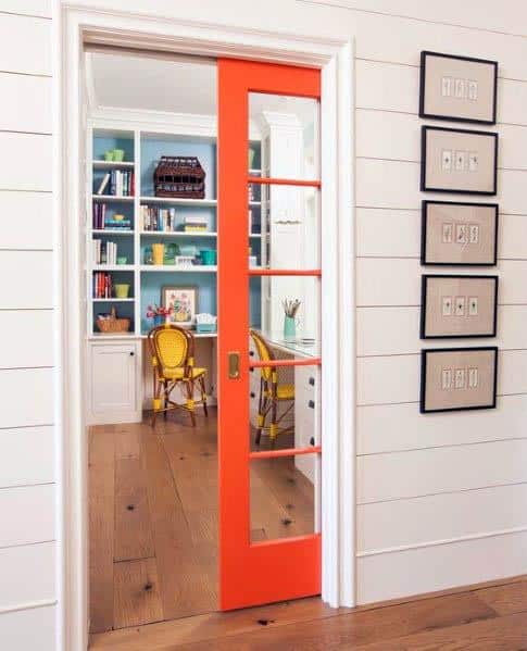 Vibrant orange pocket door with glass panels opens to a colorful home office featuring built-in blue shelves and bright yellow chairs