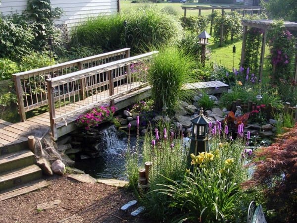 Wooden bridge over a koi pond with waterfall, surrounded by lush plants, flowers, and garden decor.