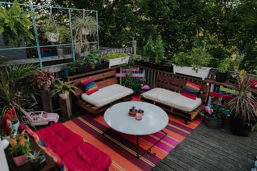 Colorful deck with DIY pallet seating, vibrant cushions, potted plants, hanging greenery, and a playful, bohemian atmosphere