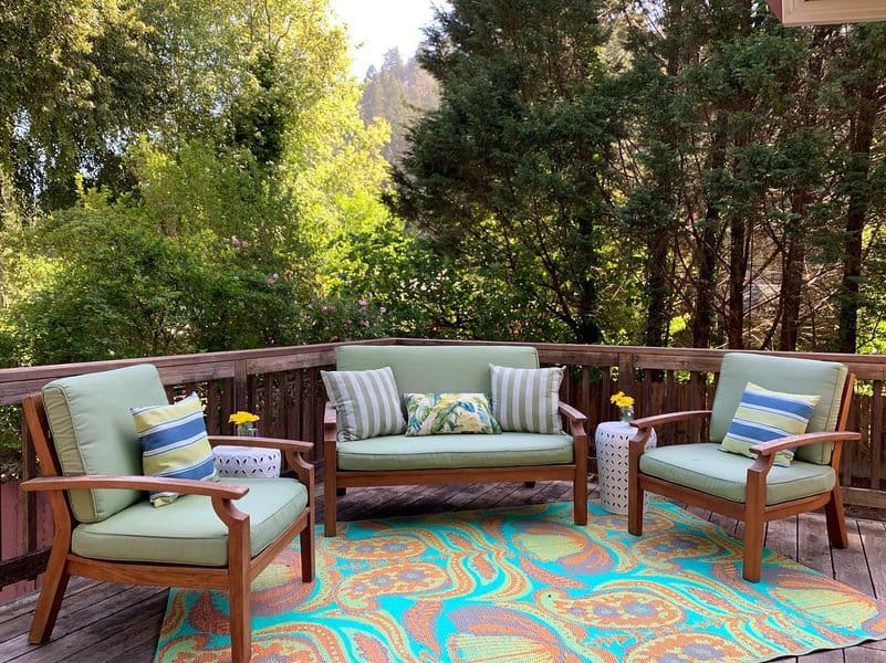 Charming deck setup featuring wooden furniture with light green cushions, striped throw pillows, a vibrant patterned rug, and lush forest views