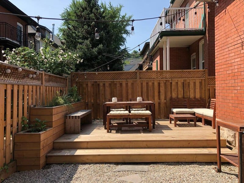 Cozy wooden deck with a bench, dining table, cushioned seating, string lights, and privacy fencing for an intimate backyard space