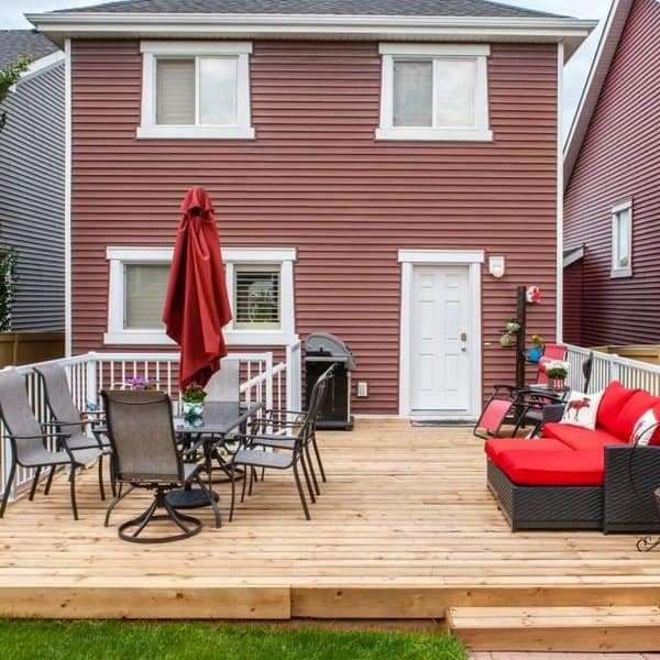 Spacious wooden deck with a dining area, red cushioned seating, a grill, and a vibrant red umbrella against a cozy home backdrop