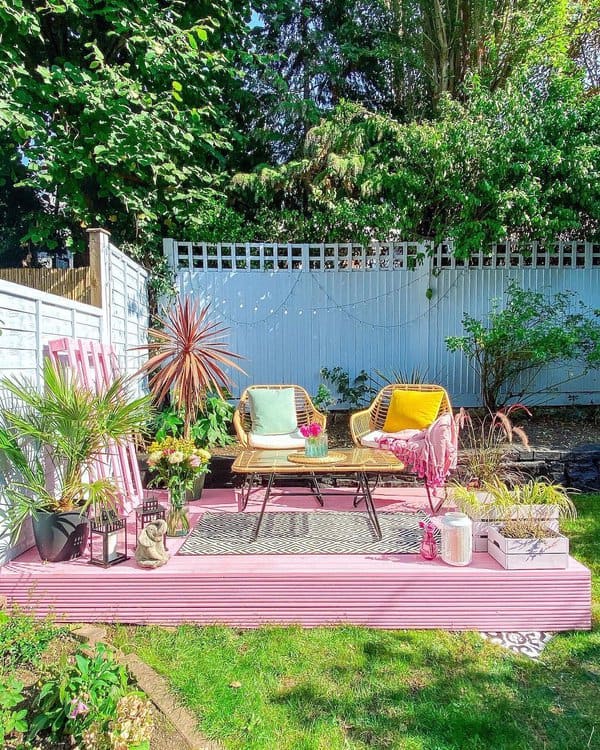 Vibrant pink deck with cozy wicker chairs, colorful cushions, potted plants, and string lights creating a cheerful garden retreat