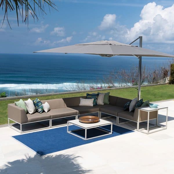 Contemporary outdoor seating area with a coastal view, featuring a neutral-toned modular sofa, vibrant accent cushions, a sleek white coffee table, and a large umbrella for shade, set on a blue rug overlooking the ocean