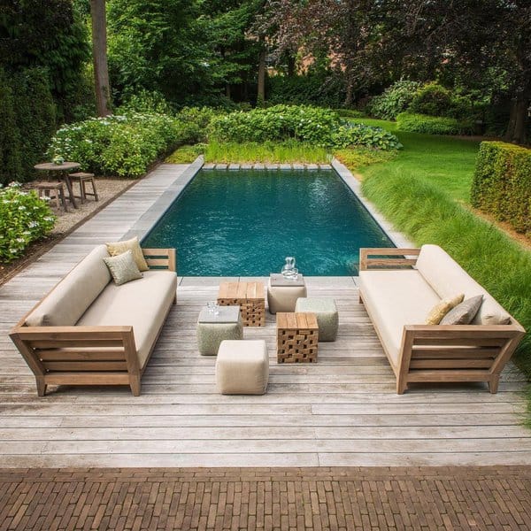 Elegant poolside deck featuring two wooden sofas with beige cushions, multiple cube-shaped tables in varying textures, and lush green landscaping creating a serene atmosphere