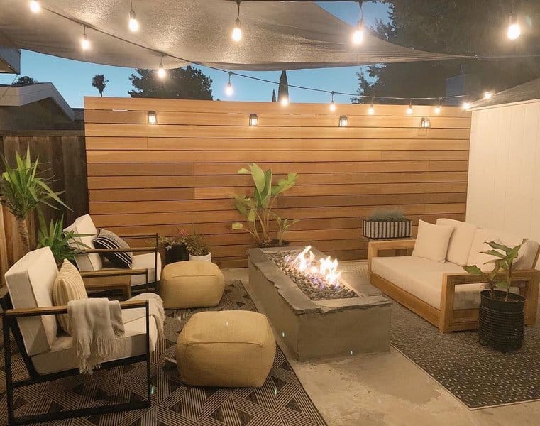 Cozy outdoor patio with a central fire pit, string lights, wooden paneling, and modern seating, creating a warm and inviting ambiance