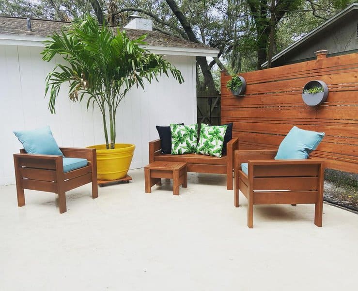 Simple and stylish DIY patio setup with wooden furniture, vibrant cushions, a large potted palm, and a modern wooden privacy fence adorned with hanging planters