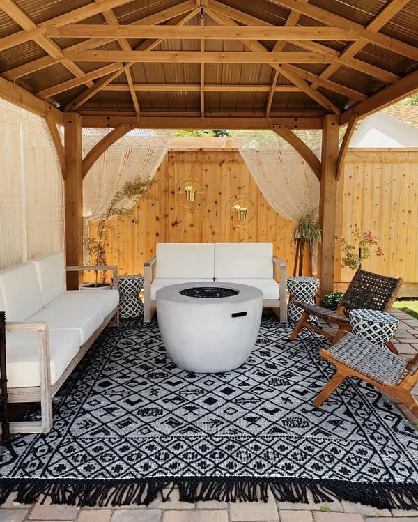 Charming pergola-covered patio with modern white seating, a central concrete fire pit, and a patterned rug for a cozy outdoor vibe