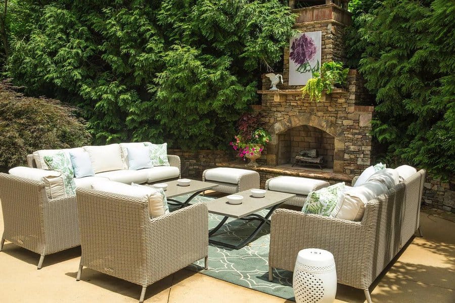 Cozy outdoor seating area with wicker furniture, a stone fireplace, and lush greenery surrounding the space