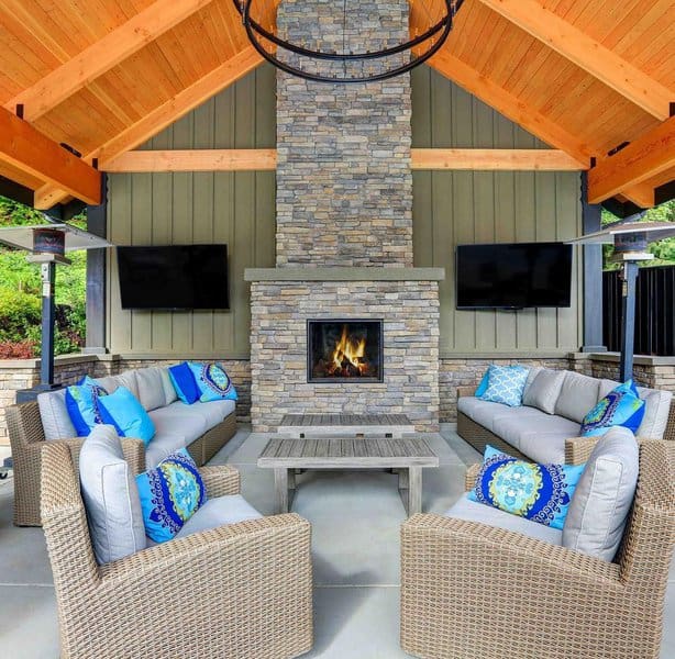 Cozy outdoor lounge setup featuring a stone fireplace under a vaulted wooden ceiling, with neutral wicker seating and vibrant blue accent pillows