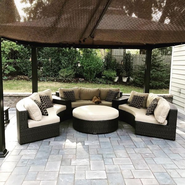 Chic outdoor lounge setup featuring curved wicker sectionals with neutral cushions, a central round ottoman, and a shaded pergola, set against a lush garden backdrop