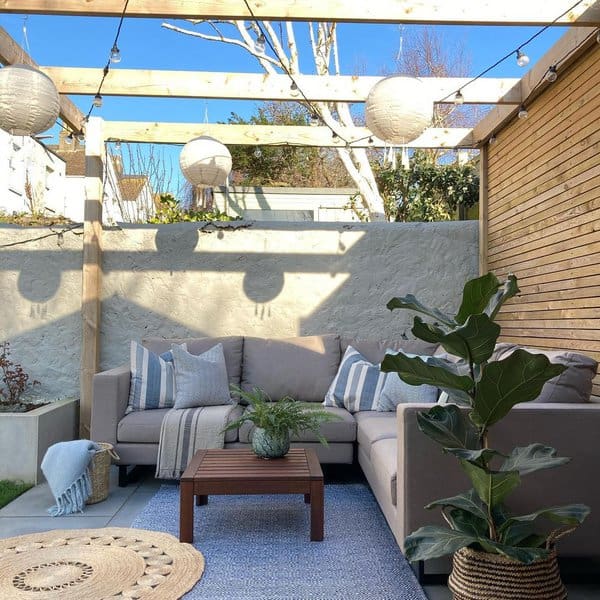 Modern outdoor space featuring a pergola with hanging lanterns and string lights, a comfortable beige sectional sofa with patterned cushions, a wooden coffee table, and lush greenery accents