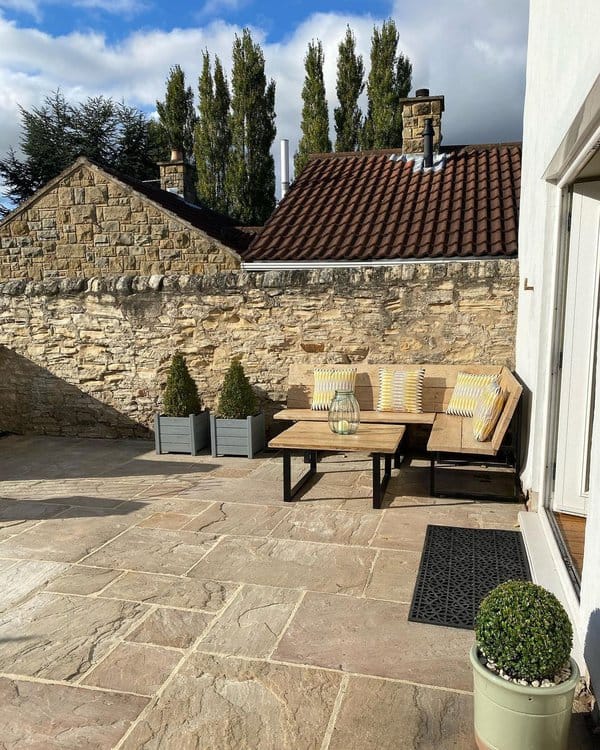 Minimalist patio with a wooden bench, striped cushions, coffee table, stone wall, and potted greenery for a cozy outdoor vibe