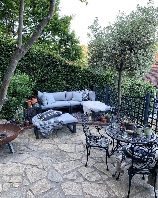 Cozy outdoor retreat featuring a stone patio, wrought-iron dining set, and a comfortable sectional with soft gray cushions, surrounded by lush greenery and potted plants for a serene garden ambiance