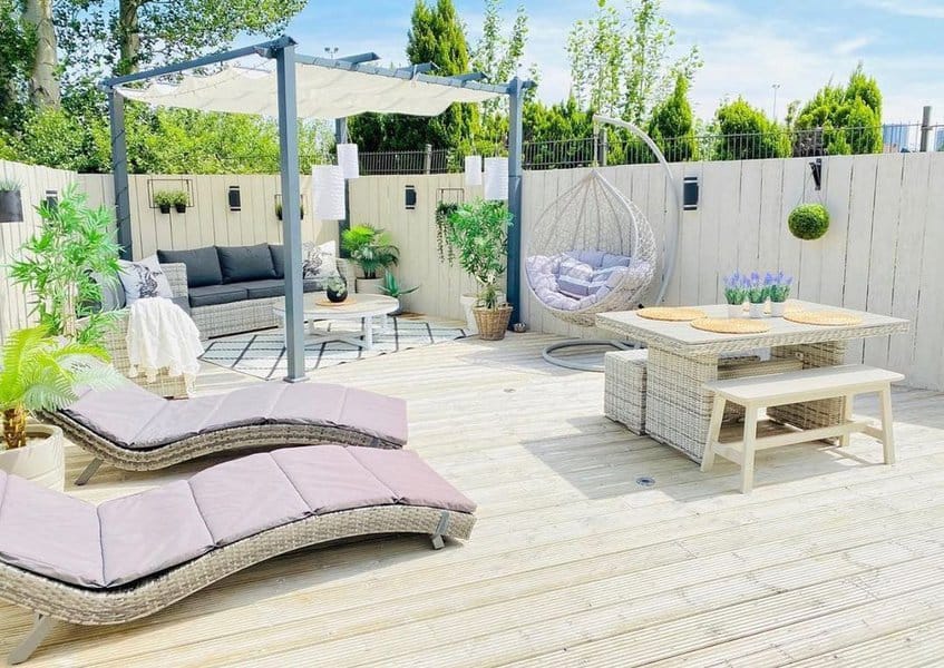 Bright and inviting outdoor space with a pergola-shaded seating area, hanging egg chair, dining bench, and sun loungers on a spacious deck, complemented by vibrant greenery and modern decor